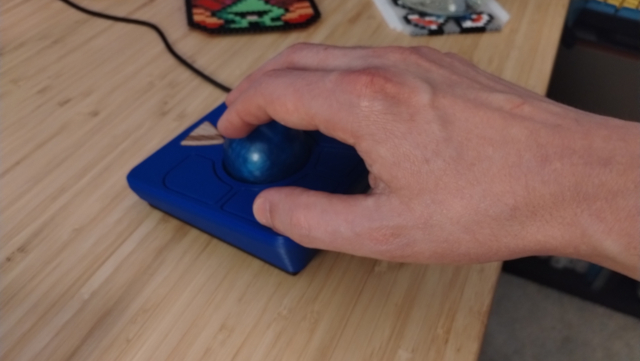 The author's hand resting on the Ploopy Adept rotated by 180 degrees and sitting on his desk. His thumb is resting on the row of buttons across the near side of the device and the tips of his fingers are resting on the trackball